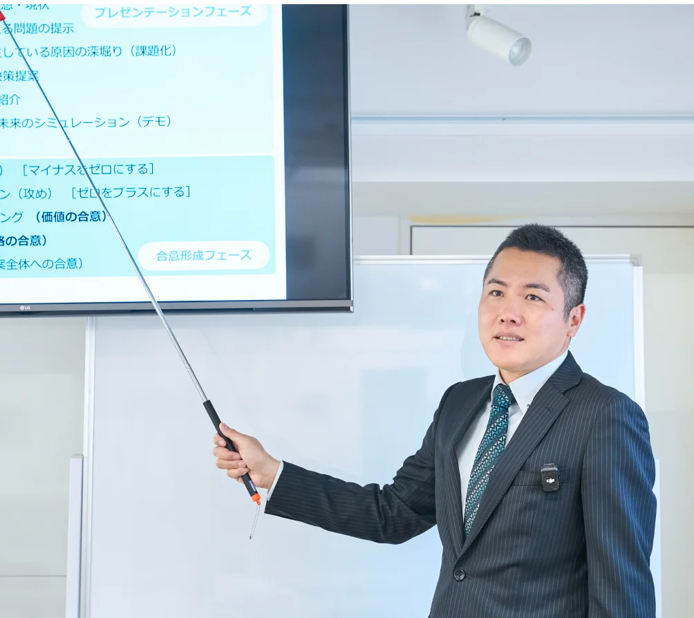 大型モニターに映された資料を指しながら説明をしている男性。